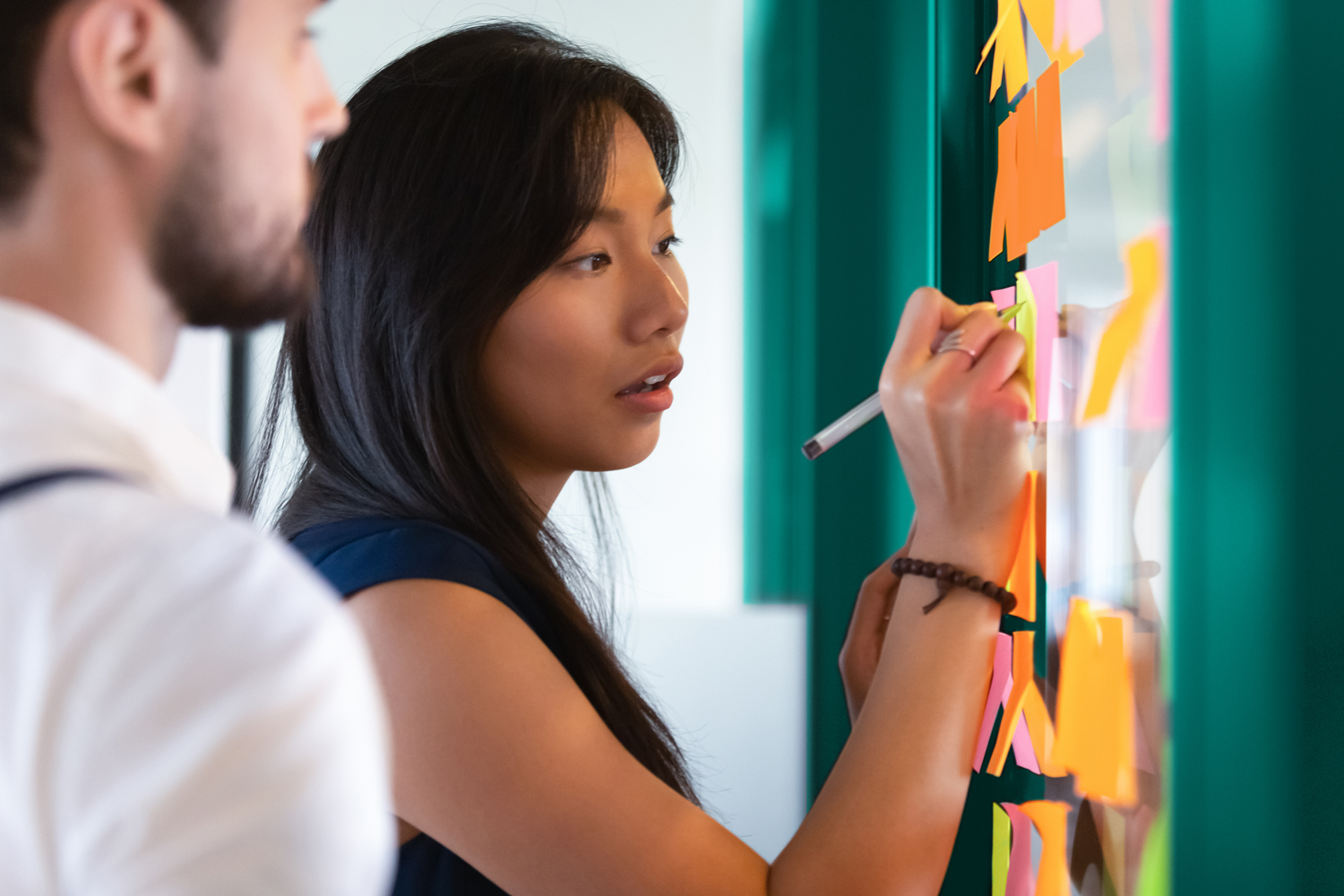 Focused asian business woman mentor coach leader writing idea or task on post it sticky notes on glass wall, serious team people developing work plan in creative corporate office at stand up meeting