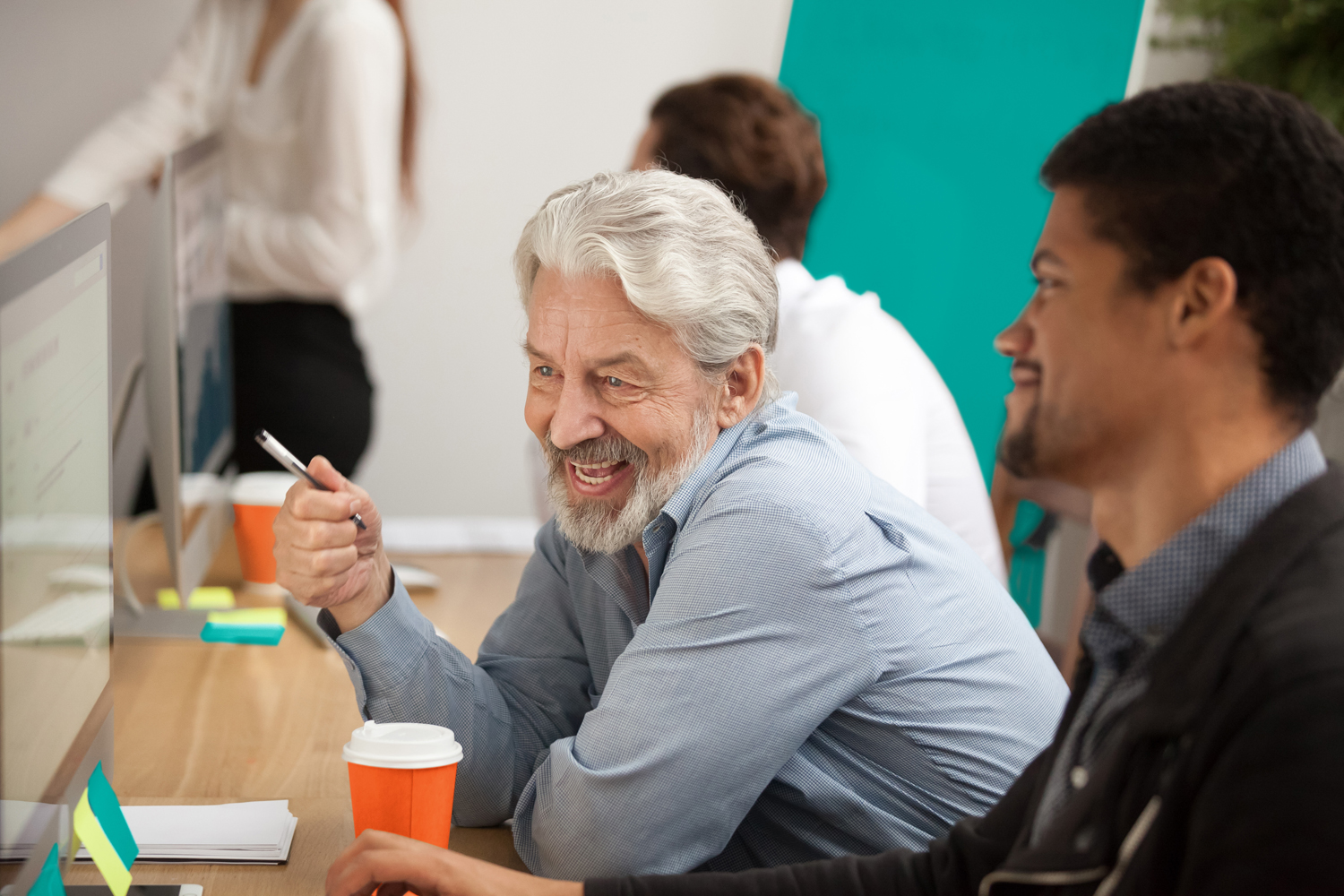 Smiling senior employee discussing email with african colleague at workplace, happy older worker talking to black coworker joking about online computer work, aged and young managers laugh in office