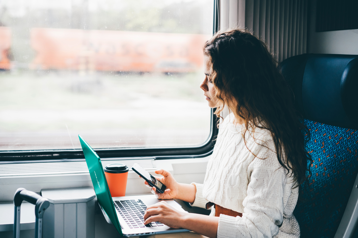 Freelancer girl working with laptop in the train. Girl looking to the phone in her hand. Business travel or technology concept.