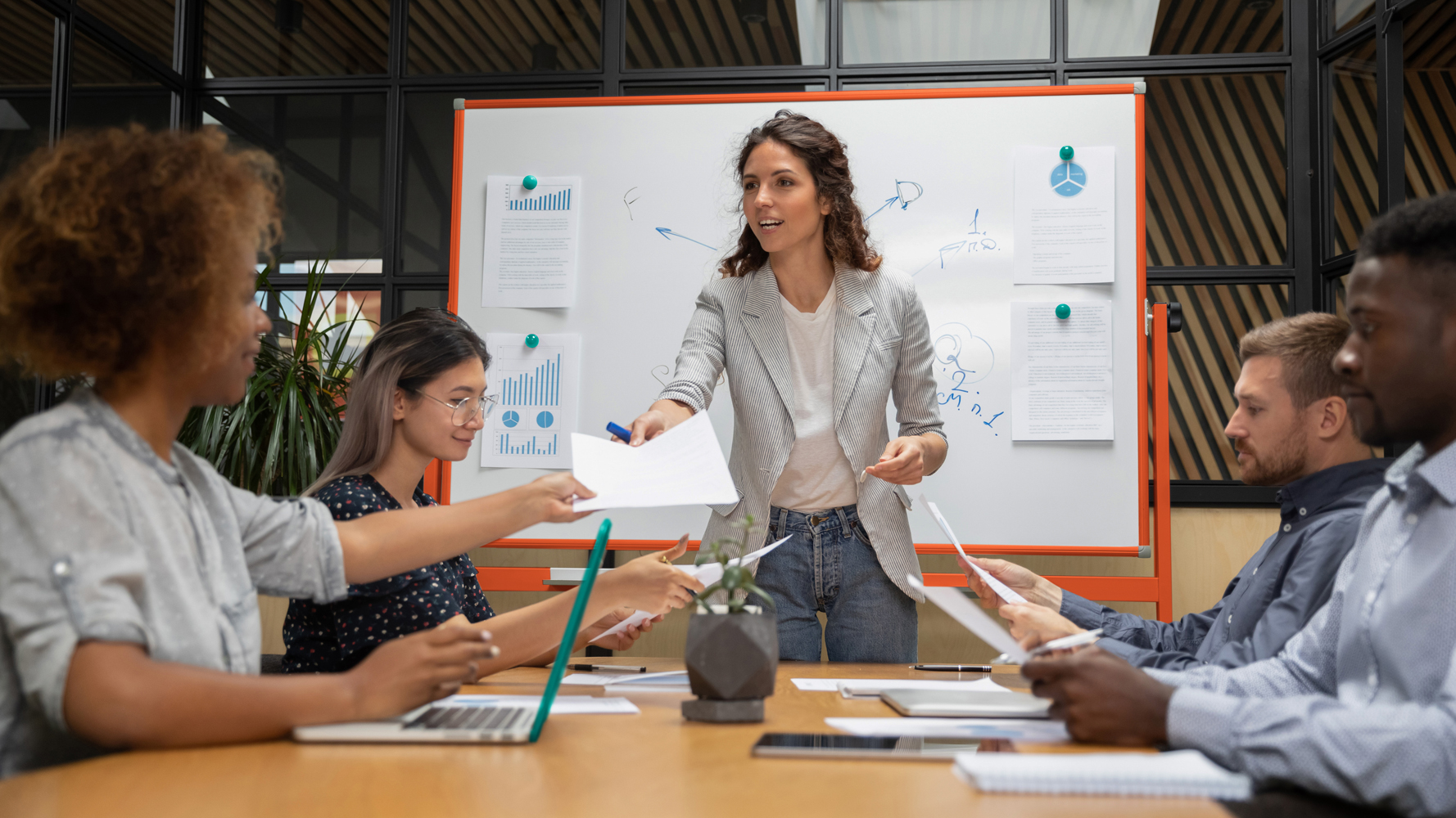 Pleasant confident millennial female boss giving out paper document report to motivated african american employee, while mixed race business people group looking through project results at meeting.