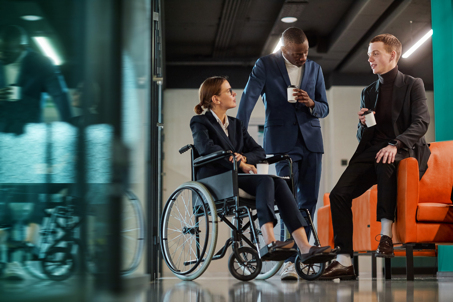 Full length portrait of diverse business team with young woman in wheelchair chatting to male colleagues in modern office